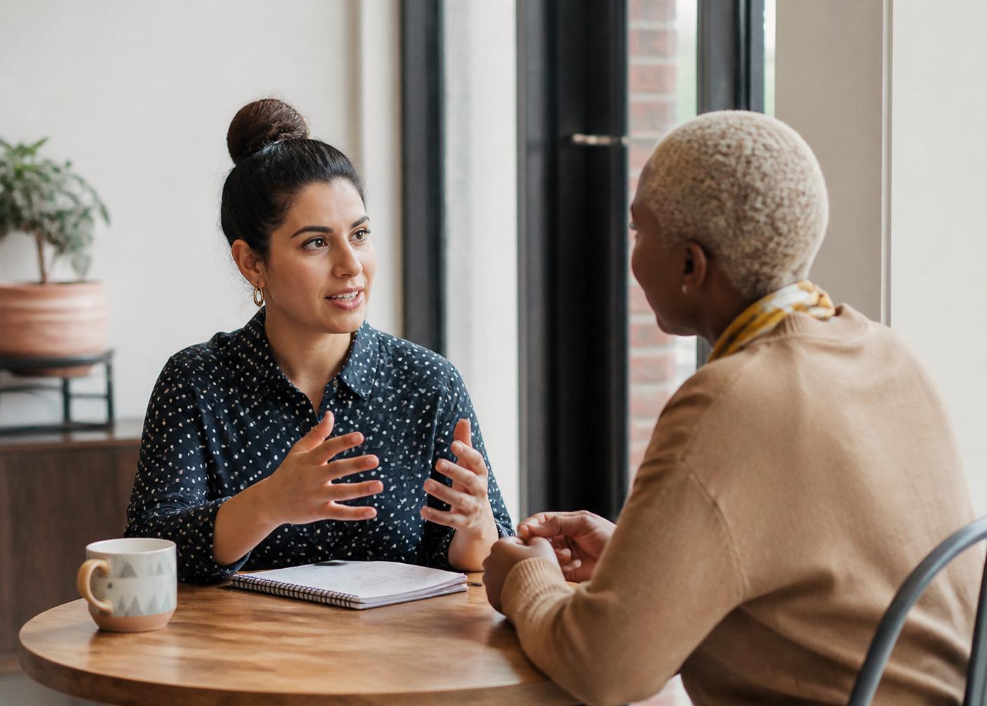 Two people in a working conversation across a small table with a notebook and coffee.